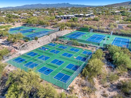 The image shows an aerial view of a complex with multiple pickleball and tennis courts surrounded by greenery and a desert landscape.