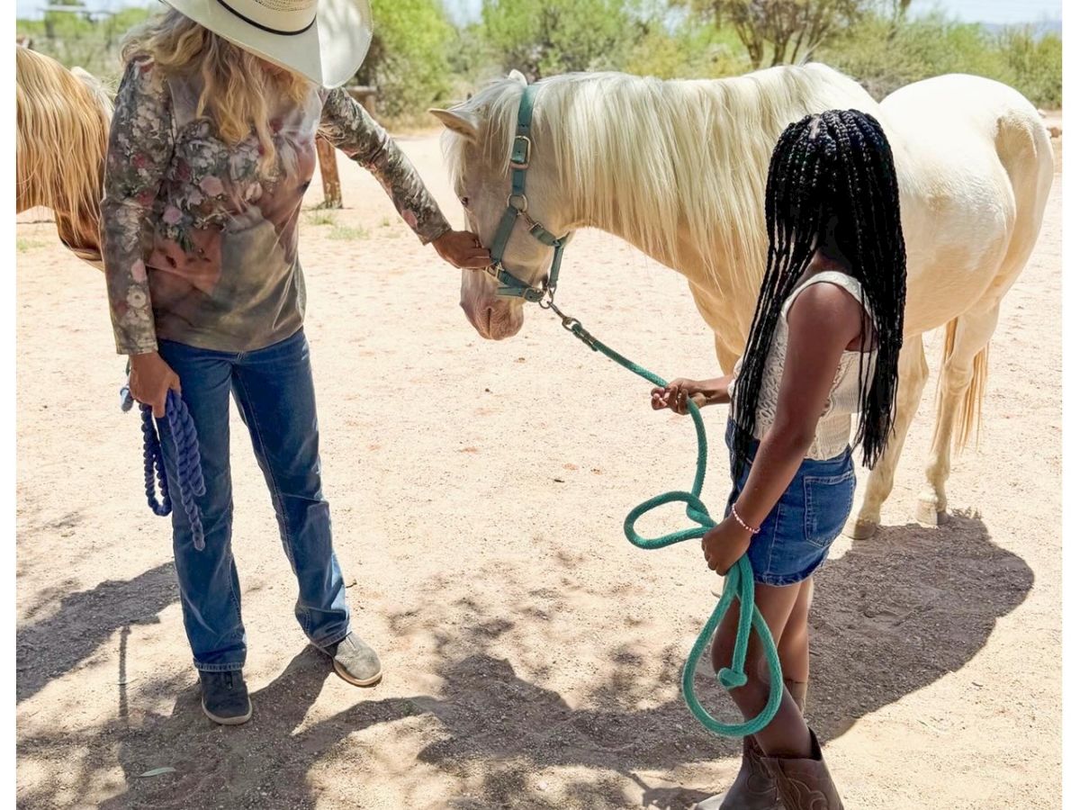 A person in a cowboy hat and another holding a rope are with a light-colored horse in an outdoor setting on a sunny day.