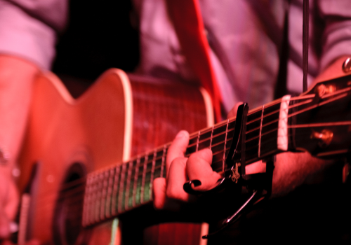 A close-up of a person playing an acoustic guitar, wearing a reddish strap and a light-colored shirt, in a dimly lit setting.