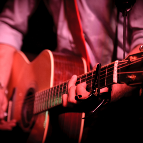 A close-up of a person playing an acoustic guitar, wearing a reddish strap and a light-colored shirt, in a dimly lit setting.