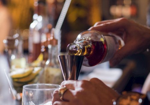 A person is pouring a drink from a bottle into a measuring jigger at a bar, surrounded by other bottles and sliced fruits, ending the sentence.