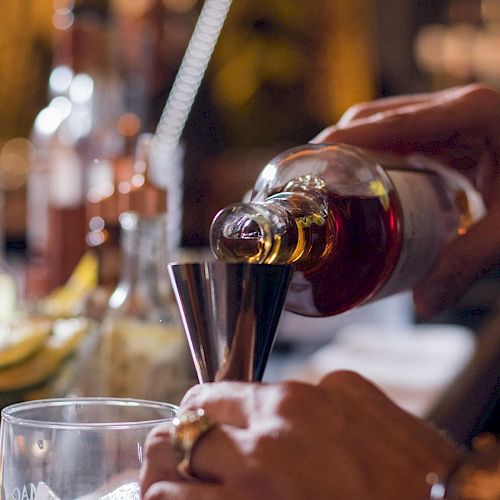 A person is pouring a drink from a bottle into a measuring jigger at a bar, surrounded by other bottles and sliced fruits, ending the sentence.