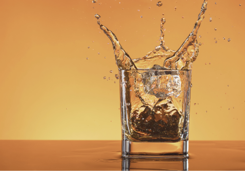 A glass with a beverage and ice cubes is in mid-splash against an orange background.