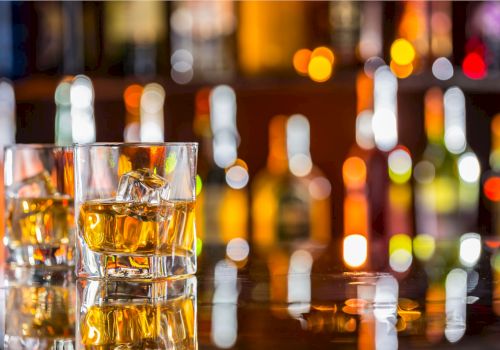 The image shows two glasses of whiskey with ice cubes on a reflective surface, set against a blurred background of colorful bottles.