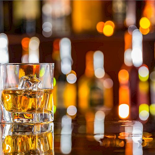 The image shows two glasses of whiskey with ice cubes on a reflective surface, set against a blurred background of colorful bottles.