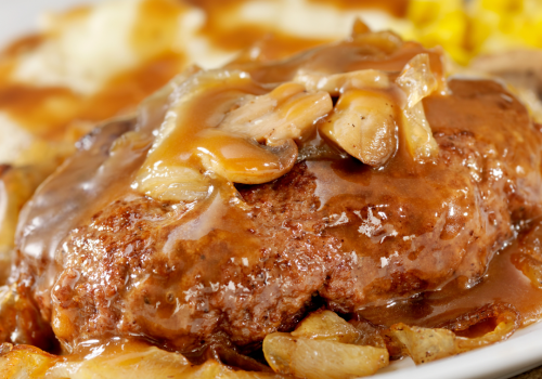 This image shows a plate of Salisbury steak covered in brown gravy with onions and mushrooms, accompanied by mashed potatoes, gravy, and some corn.