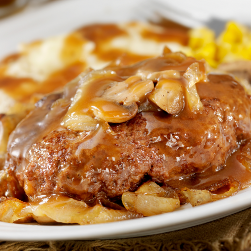 This image shows a plate of Salisbury steak covered in brown gravy with onions and mushrooms, accompanied by mashed potatoes, gravy, and some corn.