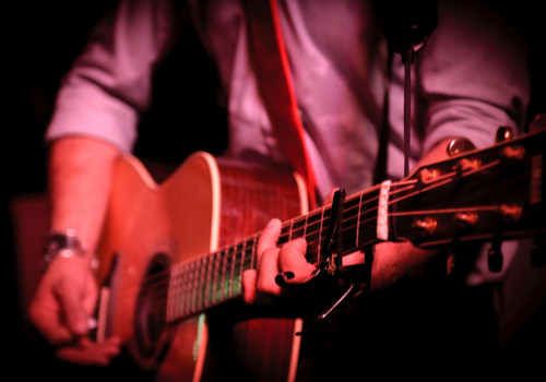 A person is playing an acoustic guitar on stage, wearing a long-sleeved shirt. The focus is on the guitar and the hands.