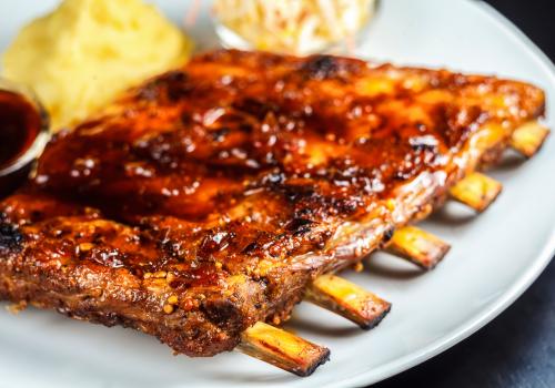 A plate of barbecue ribs with a side of mashed potatoes and coleslaw, drizzled in sauce, served on a white plate.