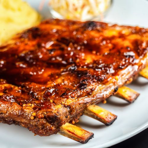 A plate of barbecue ribs with a side of mashed potatoes and coleslaw, drizzled in sauce, served on a white plate.