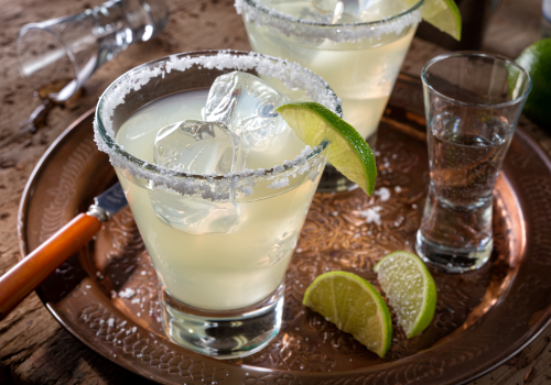 Two cocktails in salted-rim glasses with lime wedges sit on a tray, surrounded by ice, lime slices, and a shot glass.