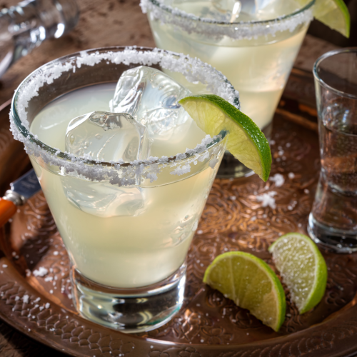 Two cocktails in salted-rim glasses with lime wedges sit on a tray, surrounded by ice, lime slices, and a shot glass.