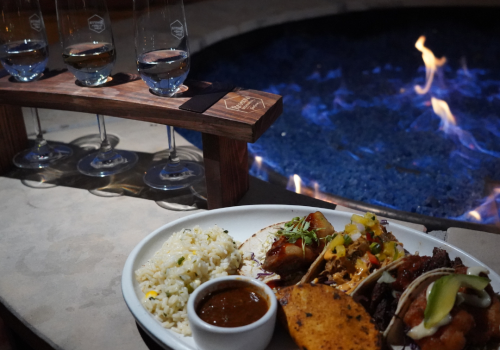 The image shows a dish with tacos, rice, and a small bowl of sauce near a fire pit, accompanied by a rack of four wine glasses on a stand.