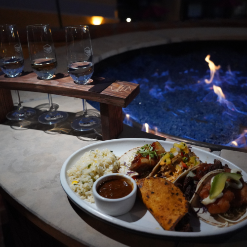 The image shows a dish with tacos, rice, and a small bowl of sauce near a fire pit, accompanied by a rack of four wine glasses on a stand.