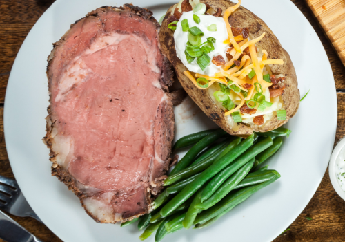 The image shows a plate with a serving of prime rib, a baked potato topped with sour cream, cheese, bacon, and green onions, alongside green beans.