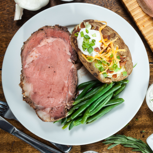 The image shows a plate with a serving of prime rib, a baked potato topped with sour cream, cheese, bacon, and green onions, alongside green beans.