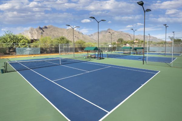 The image shows multiple outdoor tennis courts with blue and green surfaces, surrounded by desert vegetation and mountains in the background.