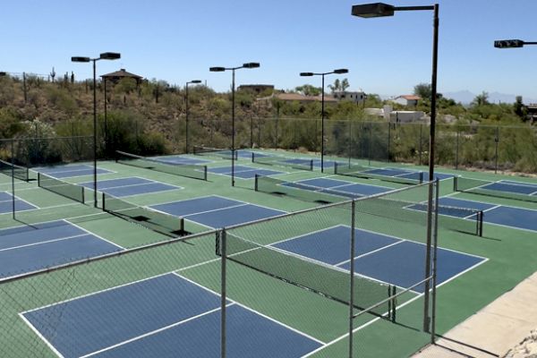 The image shows a fenced outdoor area with multiple blue and green pickleball courts. The area is well-lit with numerous tall lights.