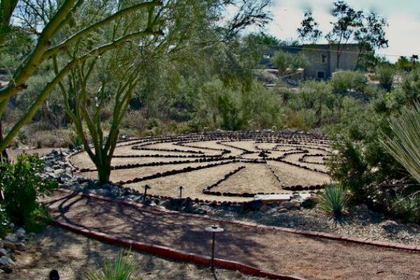 A garden scene with a labyrinth made of stones, surrounded by trees and plants, with a house in the background.