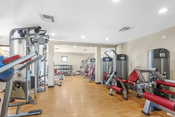 The image shows a well-equipped gym with various workout machines, including weight machines and leg press equipment, against a light-colored wall.