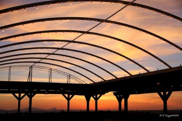 The image shows a silhouetted structure with curved beams against a backdrop of a colorful sunset, with lights hanging from the beams.