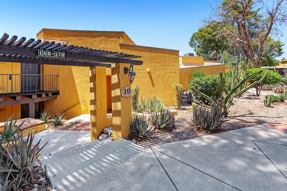 A yellow building with a pergola entrance, labeled "DEC 1948," and the number 10. The landscape includes desert plants and a concrete walkway.