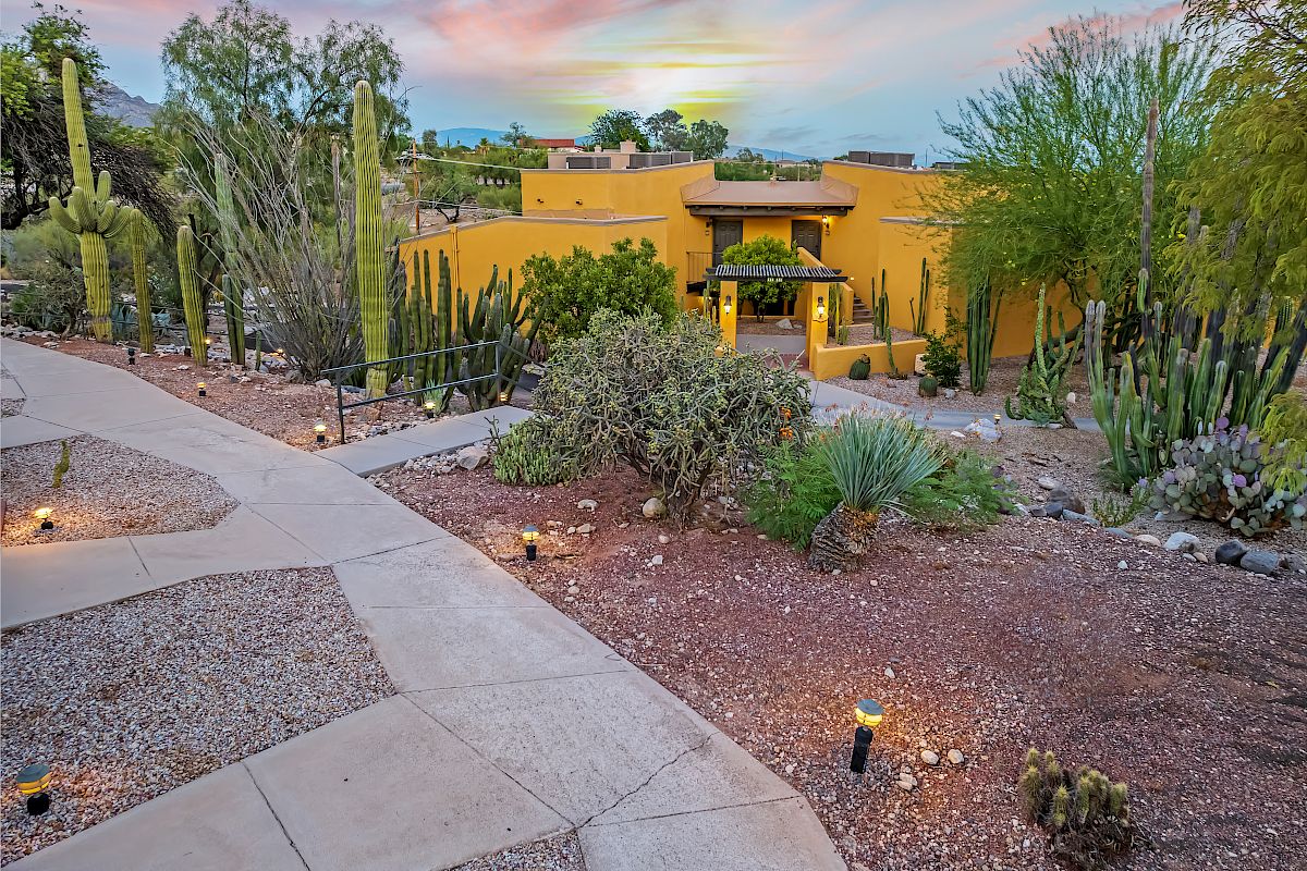 Desert landscape with cacti, pathway, and a yellow building surrounded by greenery at sunset.