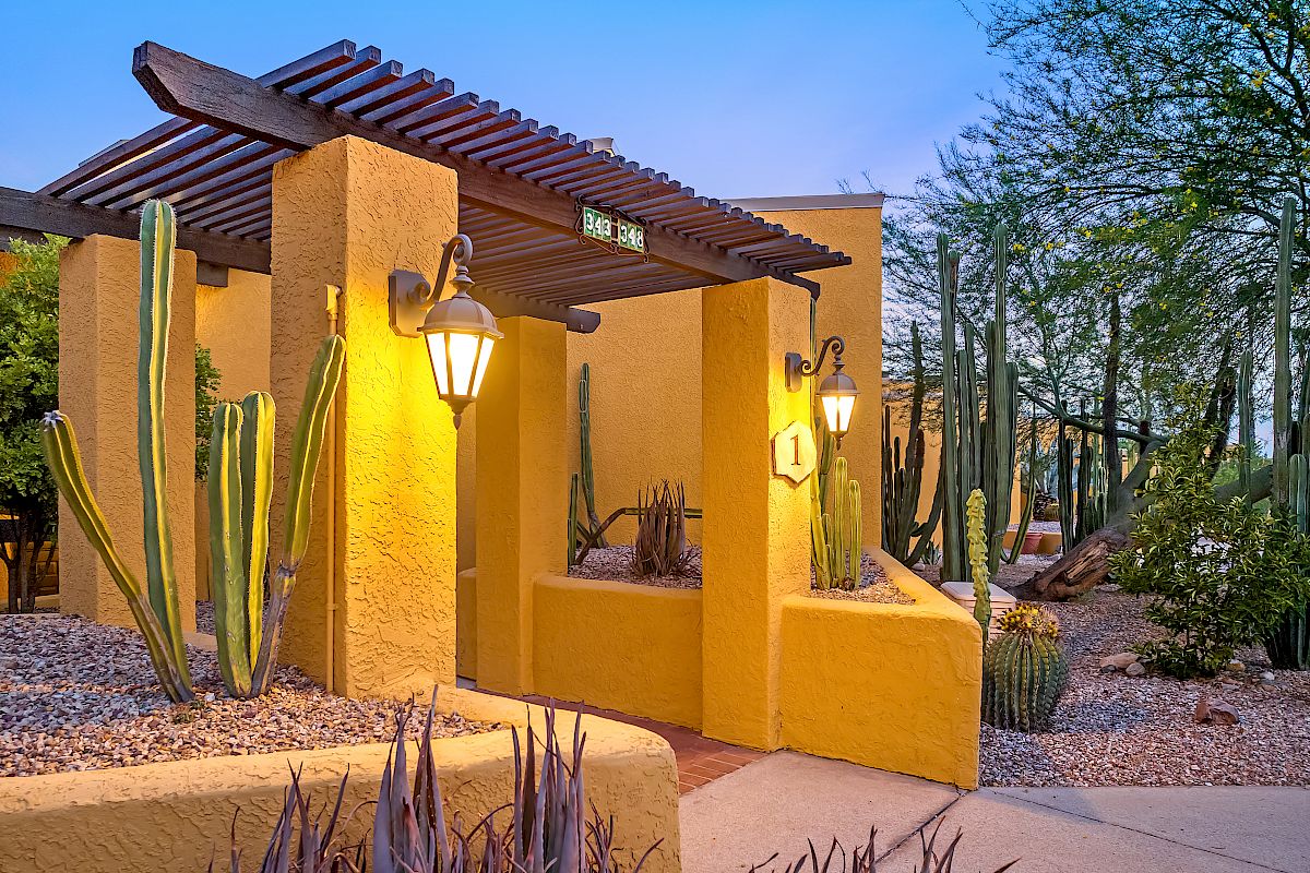 The image shows a yellow building entrance with lanterns, surrounded by cacti and other desert plants, and features a pergola at the entrance.