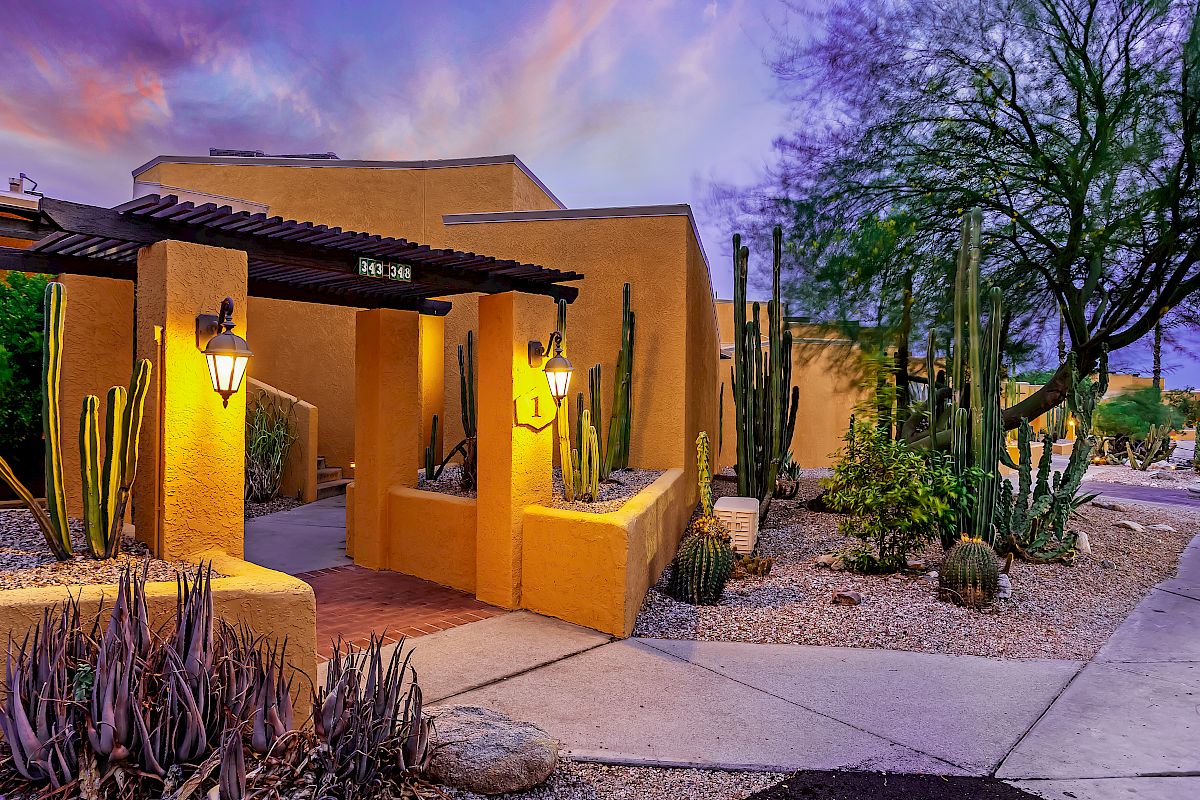 A desert-style home with a yellow stucco exterior, cacti, and lanterns illuminating the entrance path at sunset surrounded by beautiful landscaping.