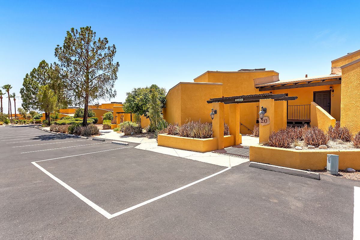 The image shows a parking lot beside modern, tan-colored buildings with lush vegetation and clear blue skies in the background.