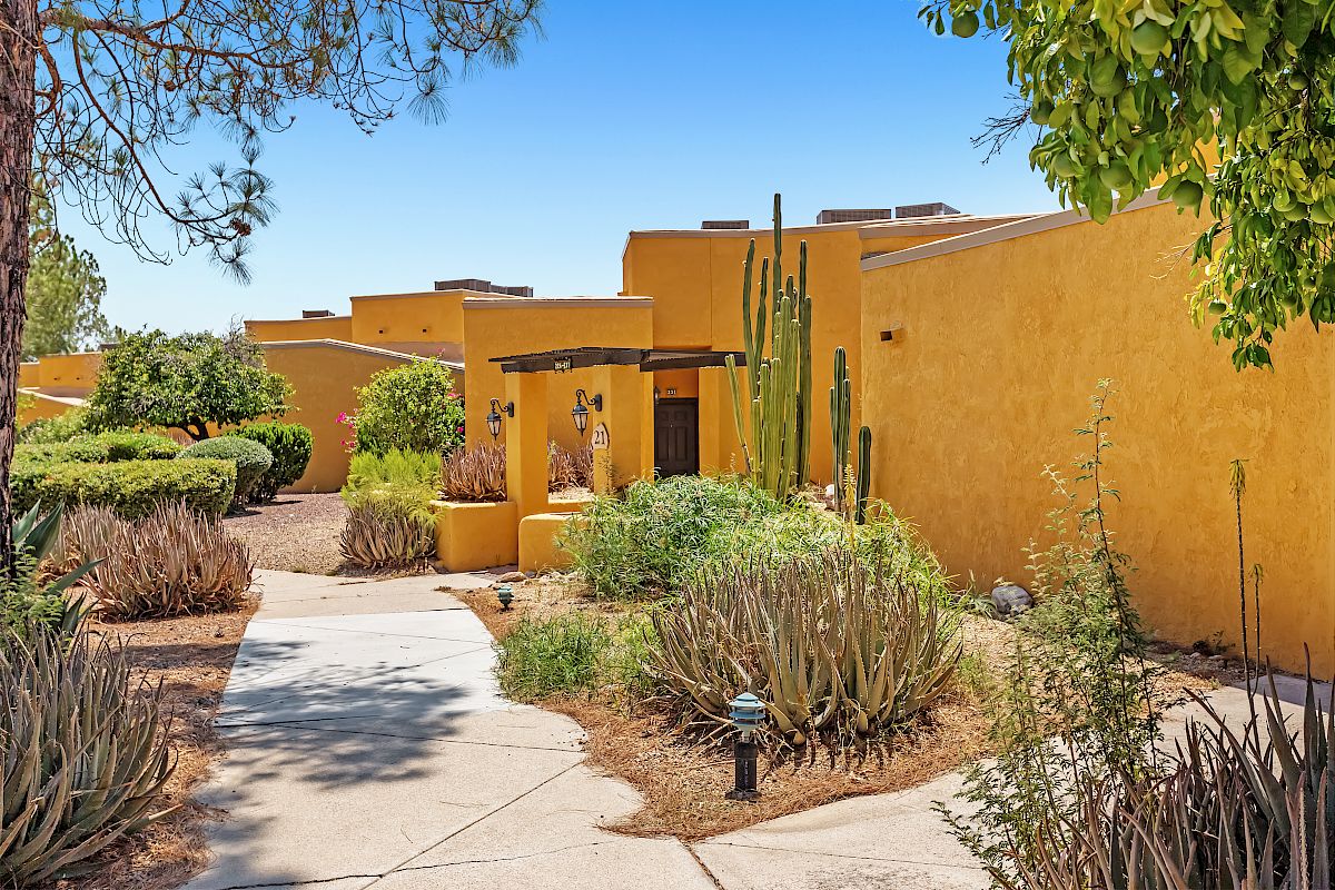 A pathway leads through desert landscaping with cactus, shrubs, and trees to a cluster of yellow adobe-style buildings under a clear blue sky.