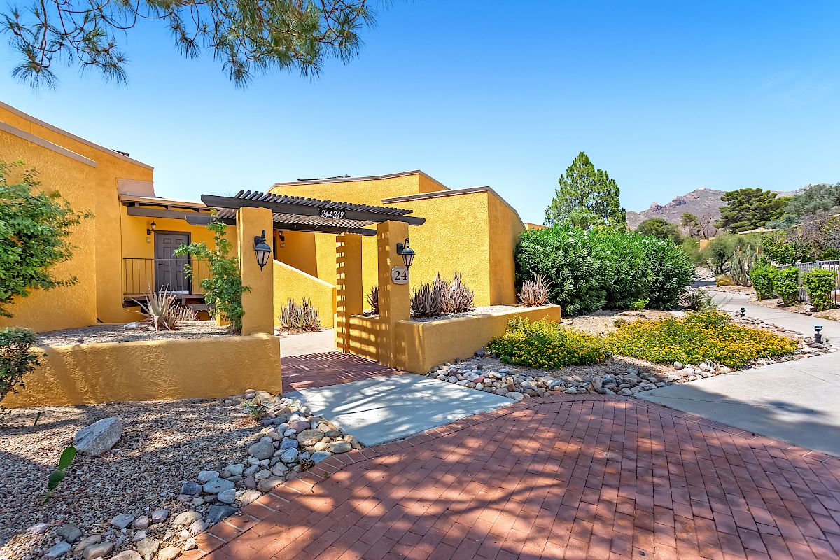 The image shows a yellow adobe-style house with a wooden pergola entrance, surrounded by desert landscaping and a brick path under a bright blue sky.