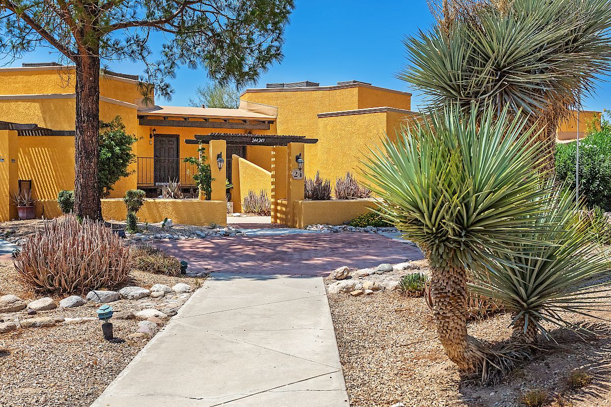 A pathway leads to a yellow adobe-style building with desert landscaping, including cacti and rocks, under a clear blue sky.