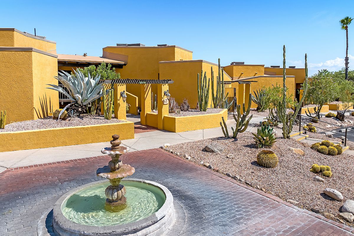 Yellow adobe-style buildings with cactus landscaping and a stone fountain in the foreground, under a clear blue sky.