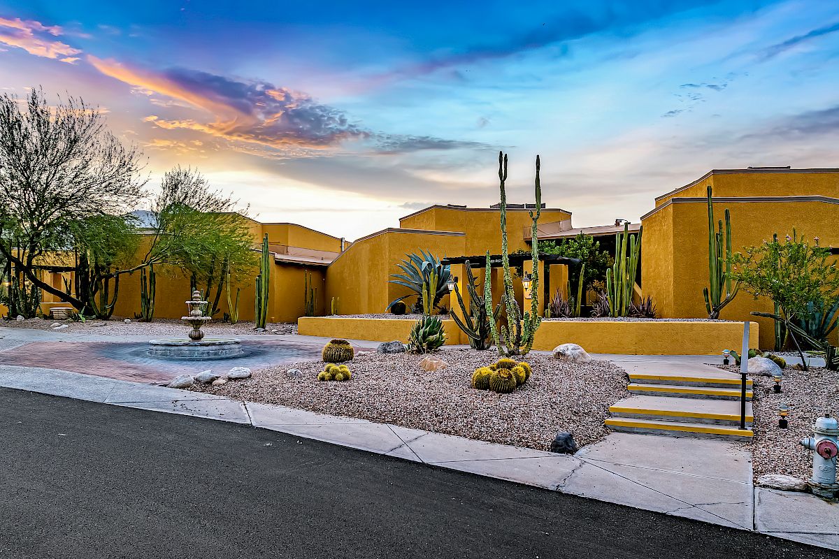 The image shows a desert-style home with a xeriscape garden featuring cacti and succulents, a paved walkway, and a fountain under a colorful sunset sky.