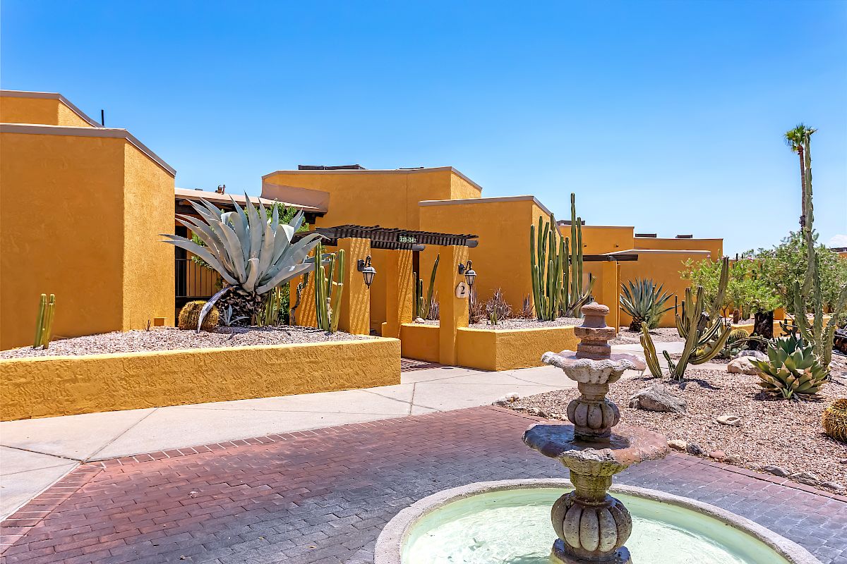 A desert-style building with yellow walls, surrounded by cacti and desert plants, with a stone fountain in the foreground, under a blue sky.
