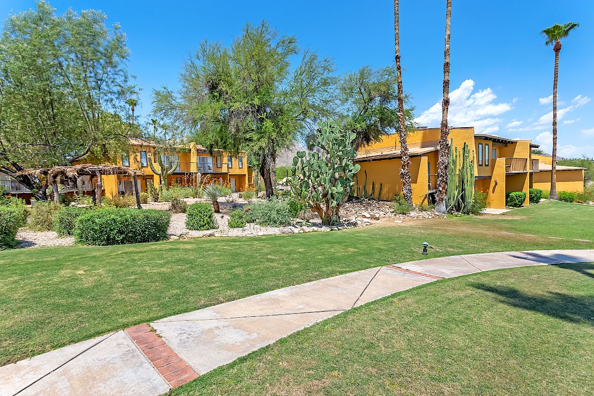 A yellow house with large windows, surrounded by trees, cacti, and landscaped lawns. There is a paved path leading to the house.