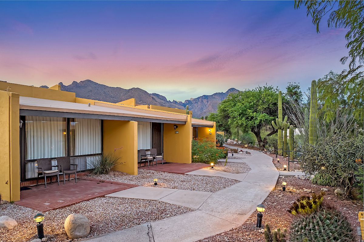 A small pathway leading to beige-colored bungalows with outdoor seating, surrounded by desert plants and lit pathway lights under a colorful sky.