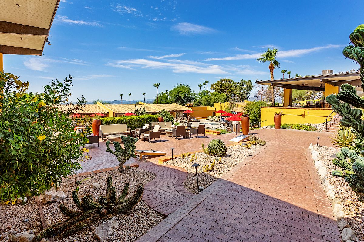 An outdoor patio area with desert landscaping, featuring cacti, seating, umbrellas, and a clear blue sky, suggesting a warm, sunny day.