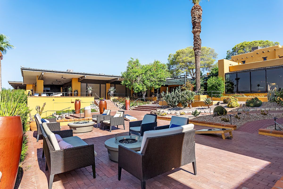 The image shows an outdoor seating area with modern furniture, surrounded by desert plants and trees, next to a large building in a sunny setting.