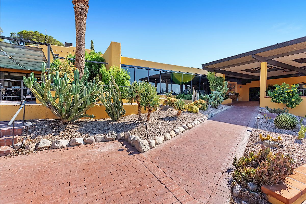 The image depicts a building with large windows, surrounded by a variety of cacti and desert plants under clear blue skies and bright sunlight.