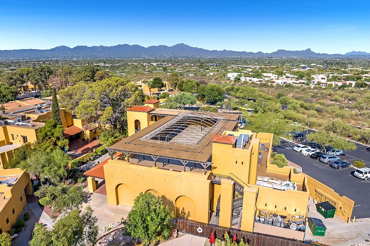 The image shows a large yellow building with arched architecture, surrounded by greenery, a parking lot, and picturesque mountains in the distance.