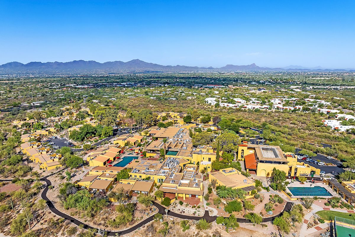 Aerial view of a sprawling desert resort complex with multiple buildings, swimming pools, and surrounding greenery, with distant mountains.