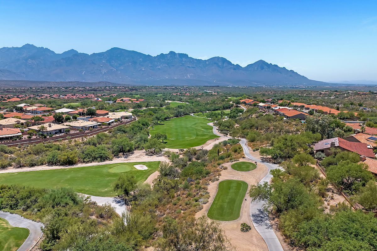 An aerial view of a golf course surrounded by greenery and residential houses with mountains in the background, under a clear blue sky.
