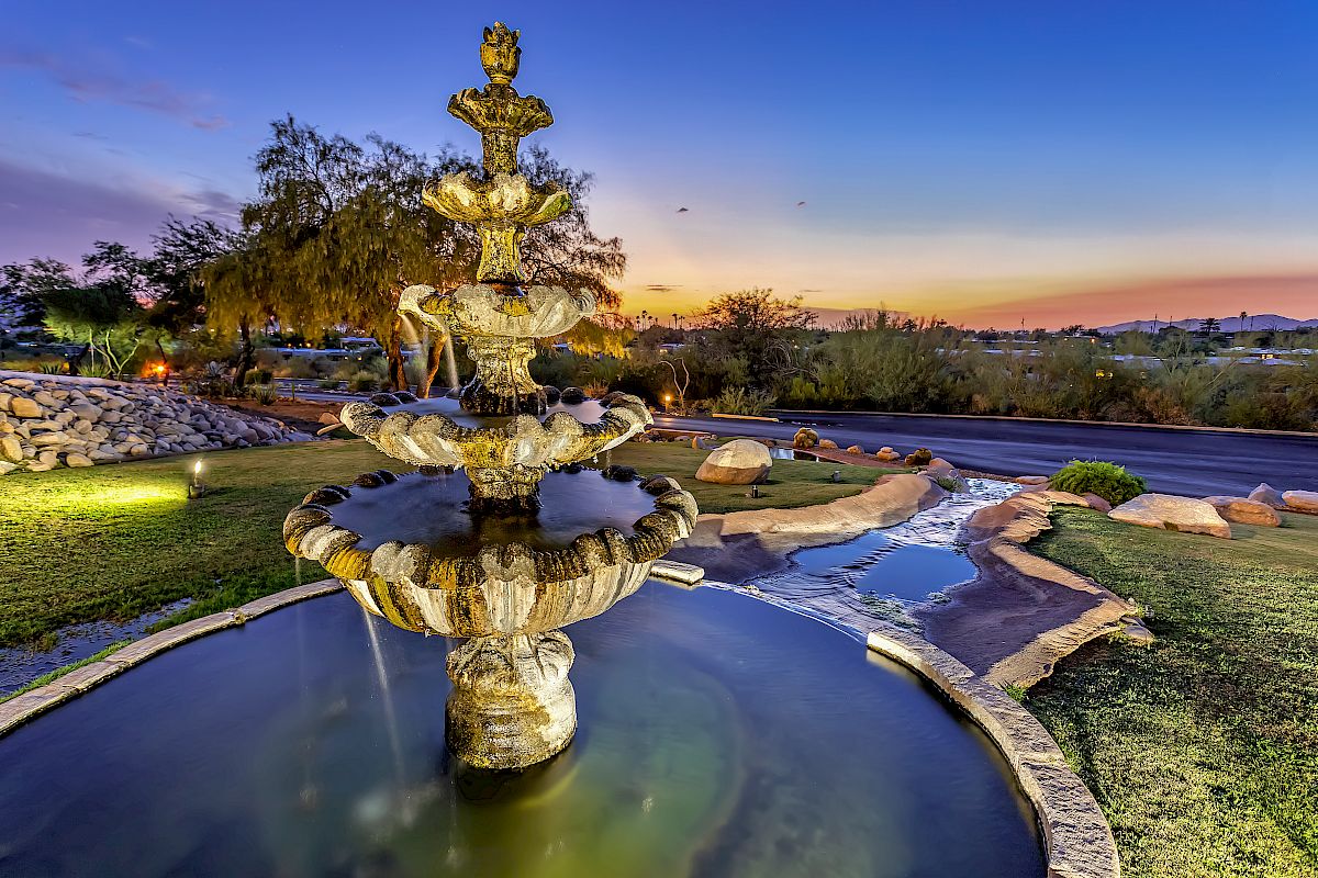 A tiered stone fountain is in the foreground, set in a landscaped park with a stream, rocks, and plants, under a dusk sky with warm lighting.