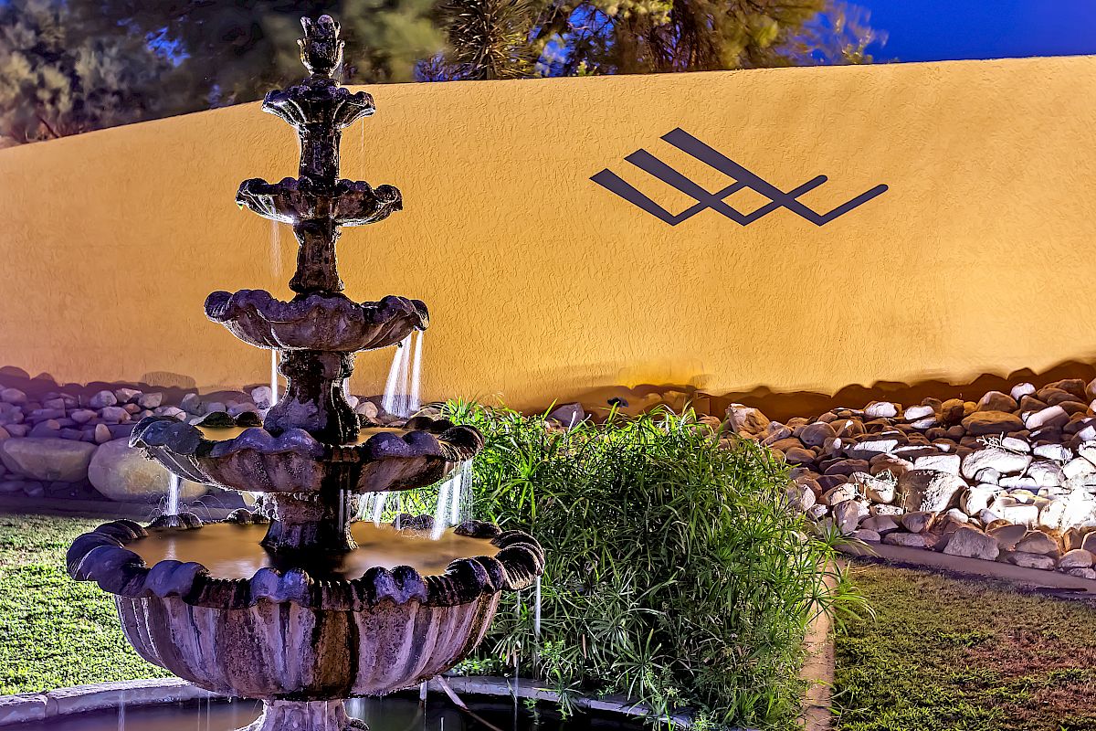 The image shows a multi-tiered water fountain in front of a yellow wall with a geometric design, surrounded by plants and rocks.
