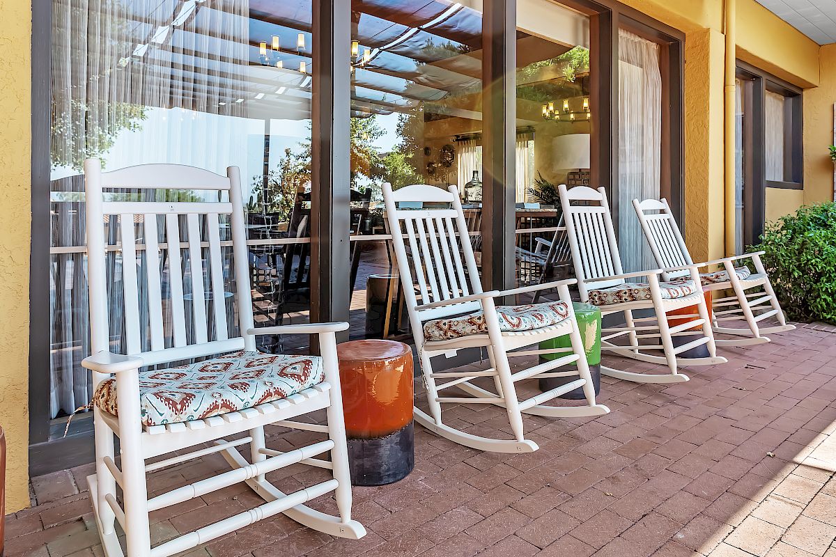 Several white rocking chairs with floral cushions on a brick patio in front of large windows, with a small side table between them.