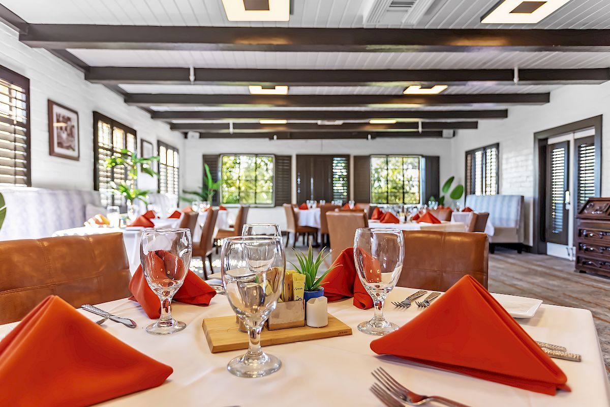 Elegant dining area with neatly set tables, orange napkins, wine glasses, and ample natural light streaming through large windows.