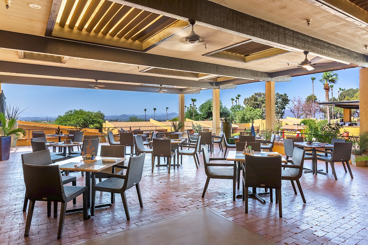 An outdoor restaurant area with several tables and chairs, a tiled floor, and a scenic view of trees and mountains in the background.