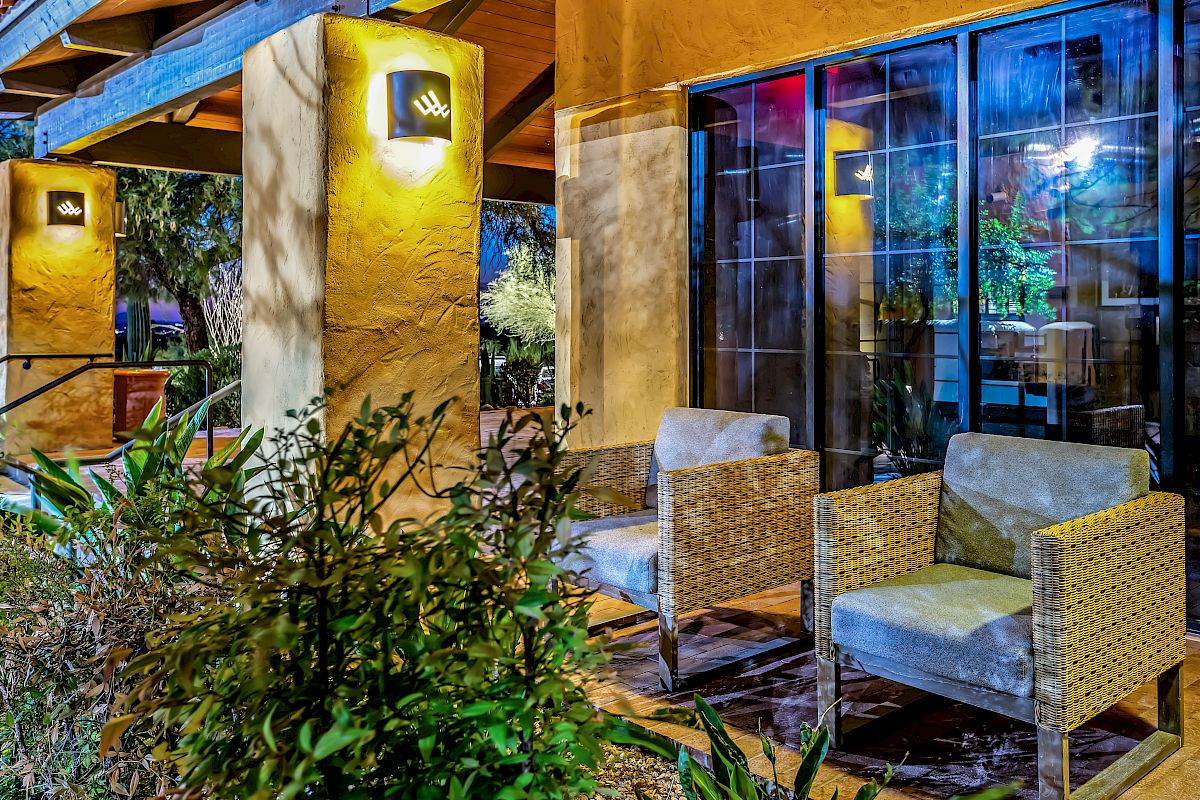 The image shows a well-lit outdoor seating area with two chairs and a lush garden in the foreground, against a backdrop of large windows.